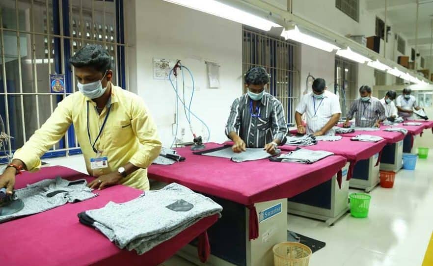 Workers in a clothing manufacturing facility ironing garments on tables with red covers.