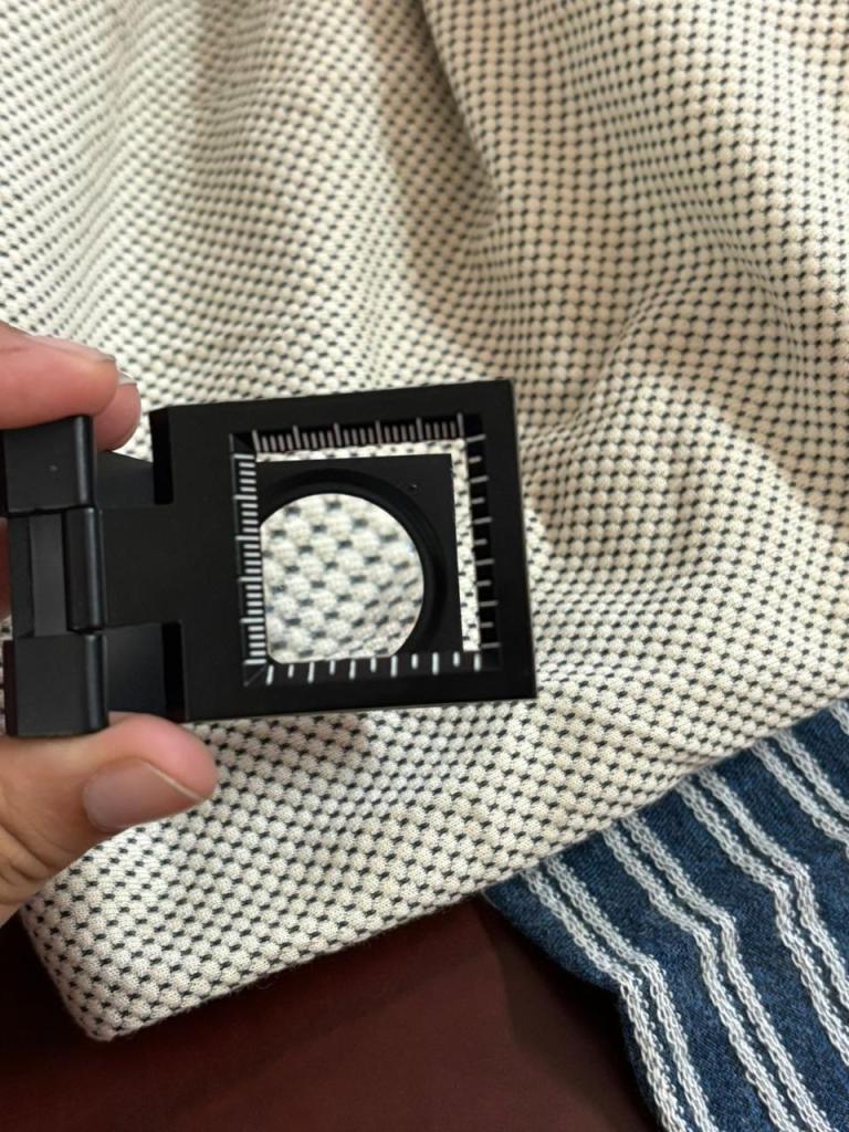 A hand holding a black measuring tool with a circular opening and a ruler, positioned over textured fabric.