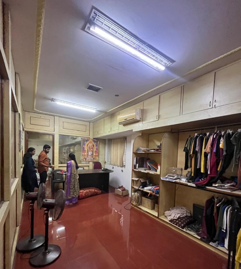 A well-lit office space with wooden cabinetry, featuring a desk, shelves of clothing, and a seating area. Three individuals are engaged in conversation, while decorative items and fans are visible in the room.