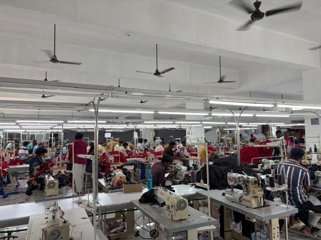 A textile workshop with multiple sewing machines and workers engaged in garment manufacturing. The space features overhead fans and bright lighting, showcasing various colorful fabrics being processed.